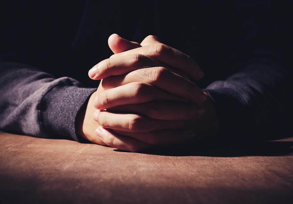 lighthousetreatment-the-complete-history-of-the-serenity-prayer-article-photo-praying-hands-of-young-man-on-a-wooden-desk-background-324611909