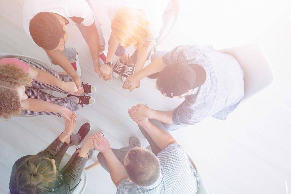 lighthousetreatment-7-questions-to-ask-if-youre-thinking-about-going-to-rehab-article-photo-high-angle-of-young-people-holding-hands-and-sitting-in-a-circle-during-group-therapy-785625016