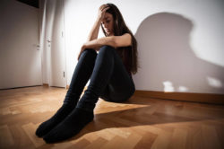 an anxious female teen sits on the floor against the white wall of her room