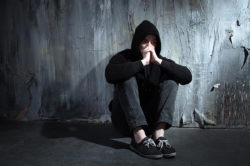 a young man in a dark hoodie sits on the floor against a black dilapidated wall