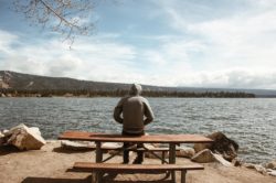 a lone man wearing a hoodie sits on a bench in front of a lake